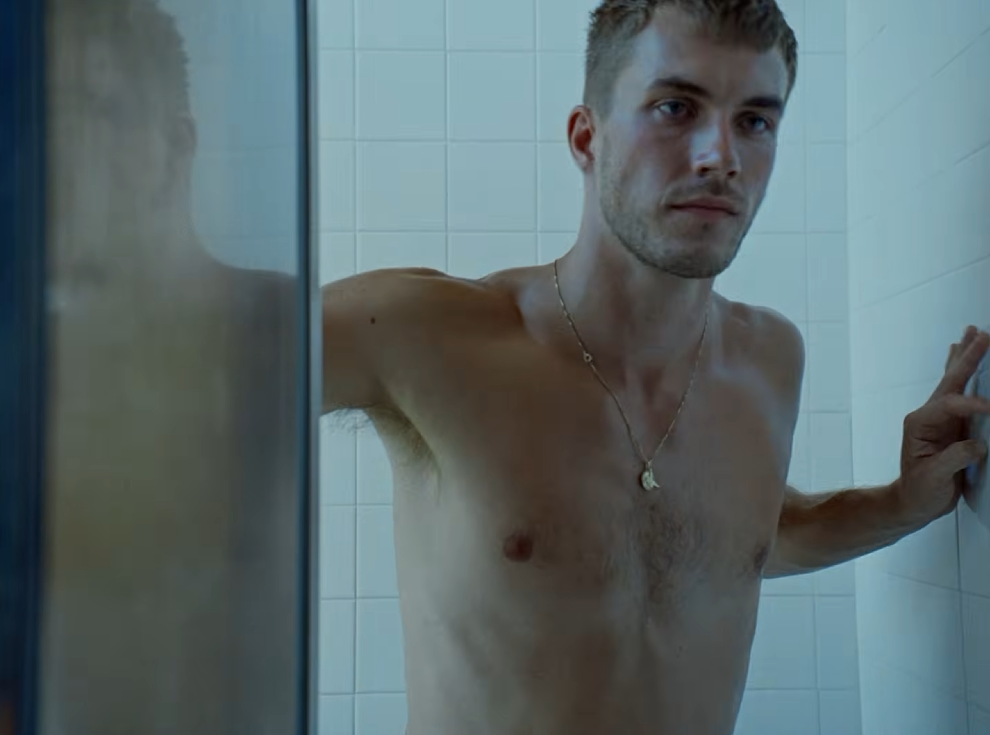 Szene aus Troye Sivans Musikvideo „FF“: Ein junger Mann mit kurzen Haaren steht mit nacktem Oberkörper in einer weiß gefliesten Dusche. Er trägt eine Halskette und stützt eine Hand an der Wand ab. Die Szene ist in kühles bläuliches Licht getaucht.