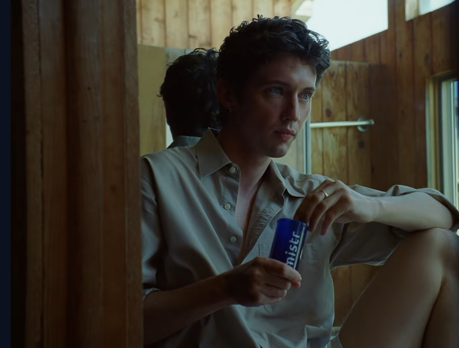 Troye Sivan sitzt in einem holzverkleideten Raum vor einem Spiegel und hält eine blaue Verpackung der HIV-Präventionsplattform MISTR in den Händen. Szene aus dem Musikvideo „FF“.
