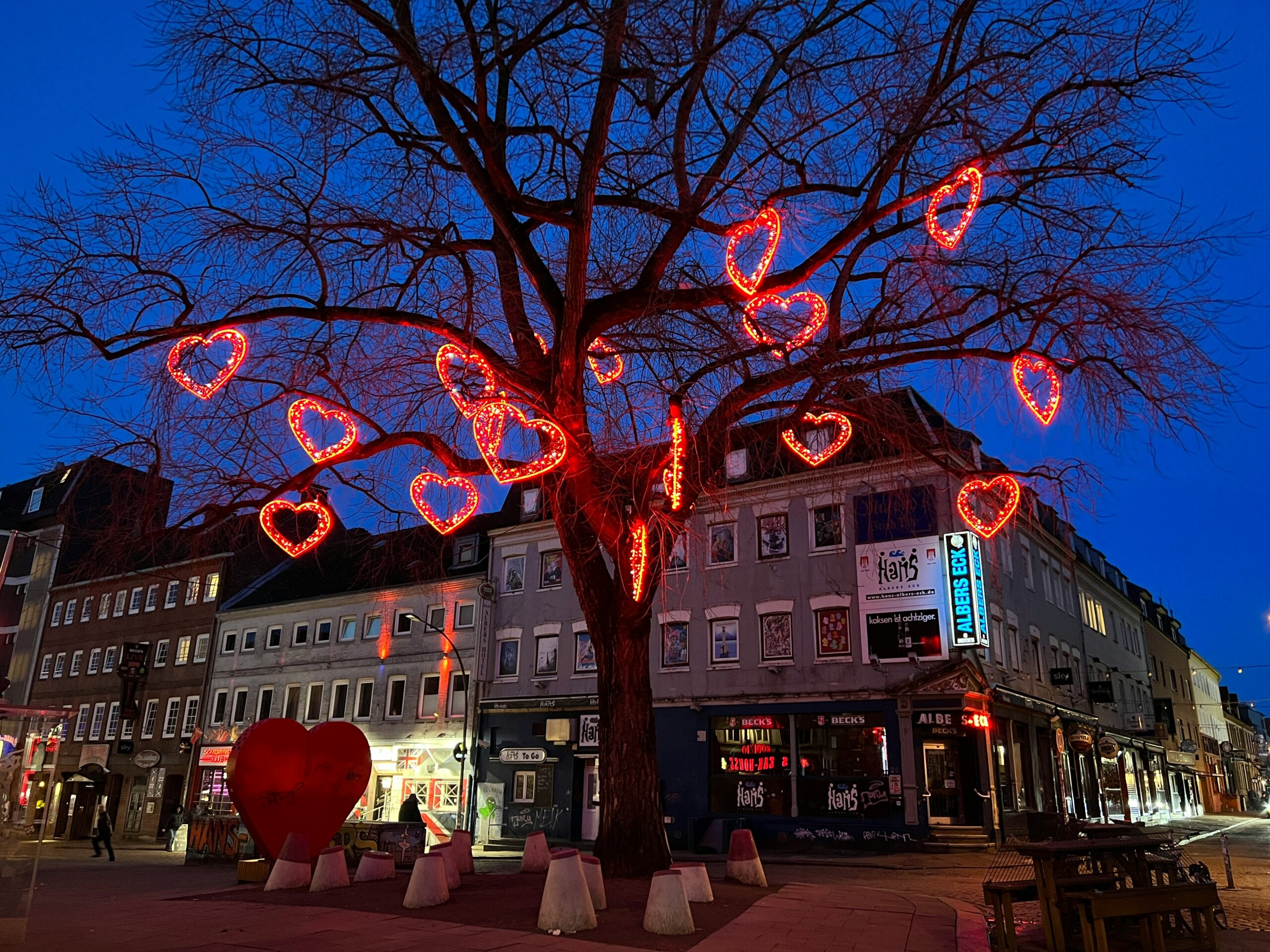 Ein kahler Baum am Hans-Albers-Platz auf St. Pauli in Hamburg, geschmückt mit rot leuchtenden Herzen als Winterdekoration. Im Hintergrund sind Kneipen und Häuser im Abendlicht zu sehen.